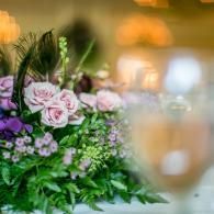 Pink roses and purple flowers in a bouquet with a blurred wine glass on a warm, glowing table setting