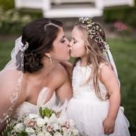 Bride kissing young girl in white dress, both holding bouquet at an outdoor wedding