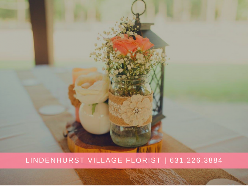 Flower arrangement in a glass vase with pink roses and greenery on a table, Lindenhurst Village Florist sign below