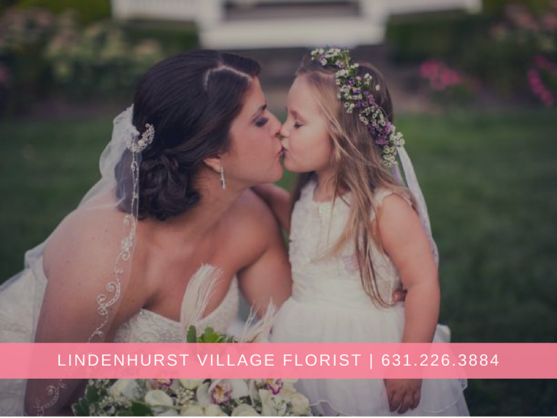 Bride kissing young girl in white dress, with floral crown, at outdoor wedding; Lindenhurst Village Florist banner below