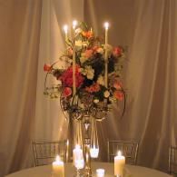 Candlelit floral centerpiece on a table with tall candles and draped curtains in the background