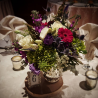 Floral centerpiece with purple, pink, and white flowers on a table set with candles and folded napkins