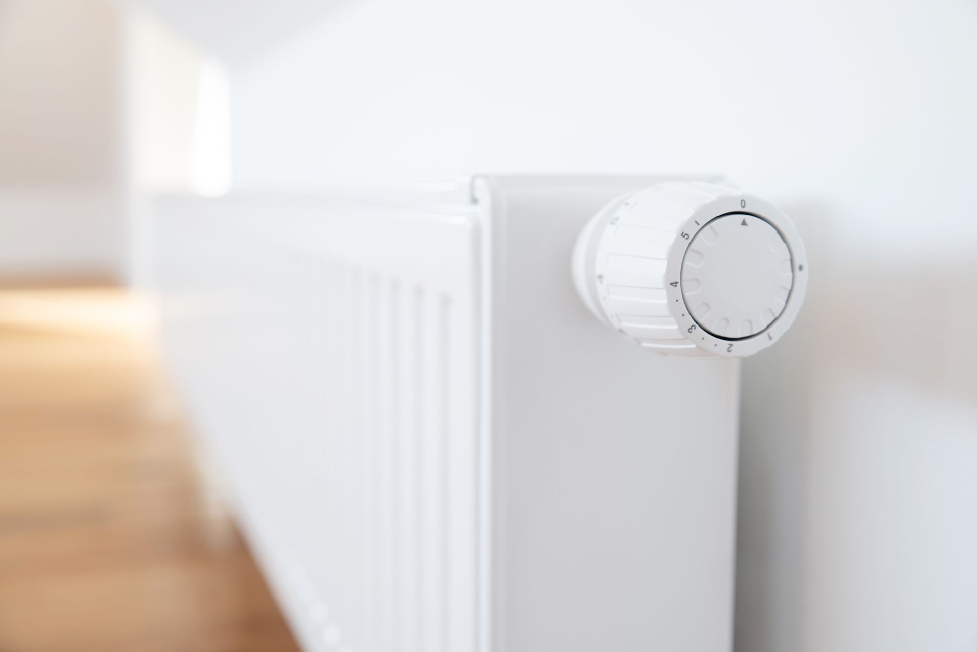 Weißer Heizkörper mit Temperaturregler vor einer weißen Wand in einem Raum mit Holzboden.