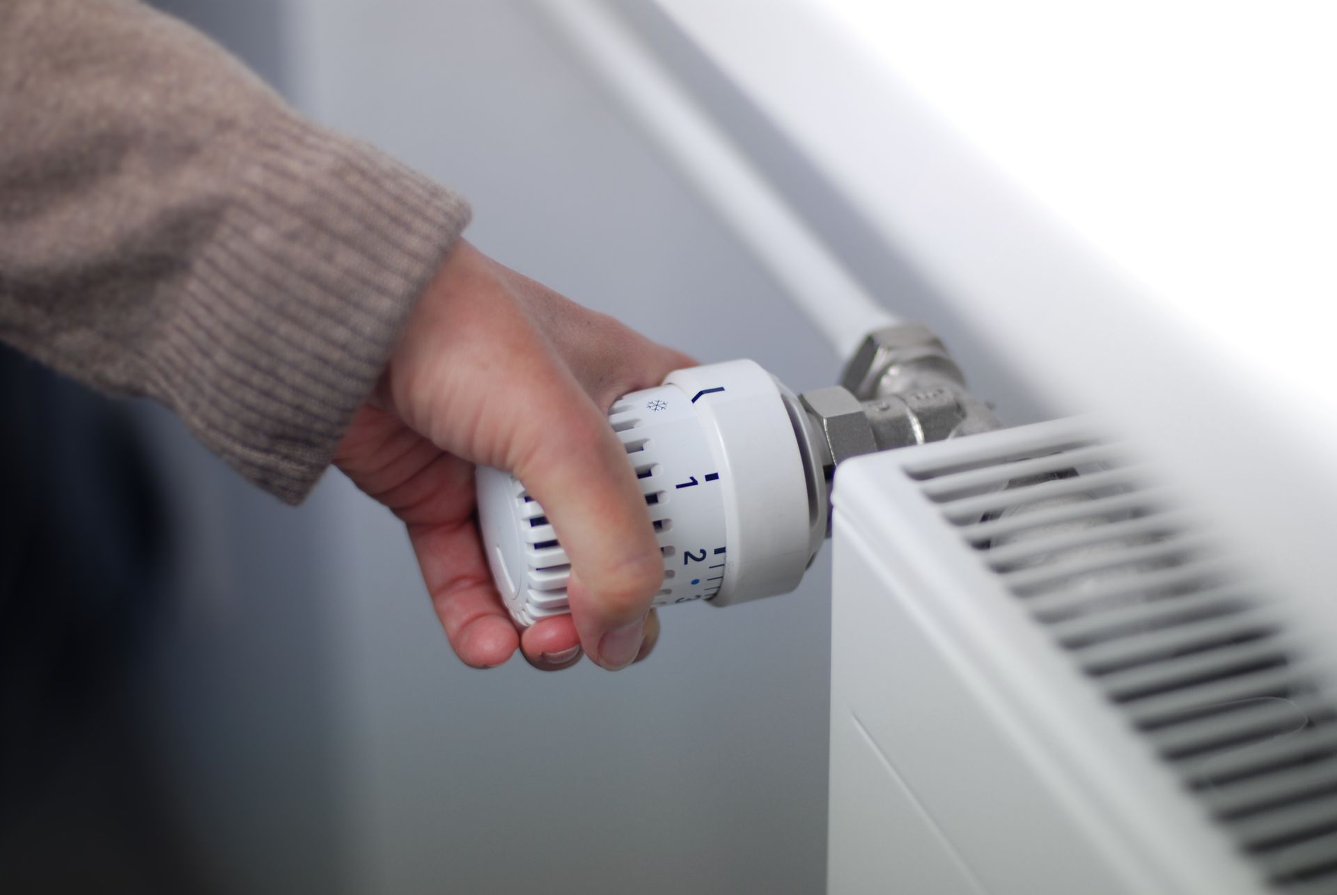 Eine Hand dreht an einem weißen Thermostatknopf an einem Heizkörper.