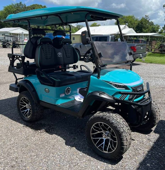 Turquoise off-road golf cart with raised tires parked on gravel under a canopy