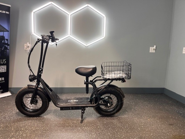 Black electric scooter with rear basket parked indoors against a gray wall with hexagon lights.