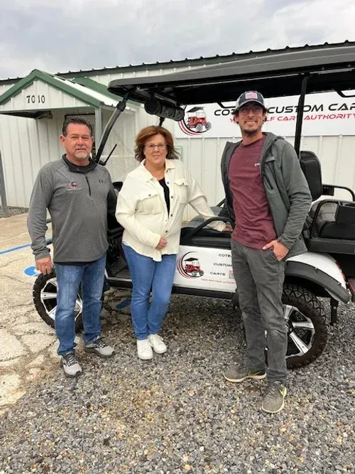 Three people standing beside a white golf cart under a canopy outdoors.