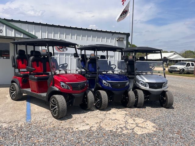 Three parked golf carts in red, blue, and silver outside a metal building on gravel.