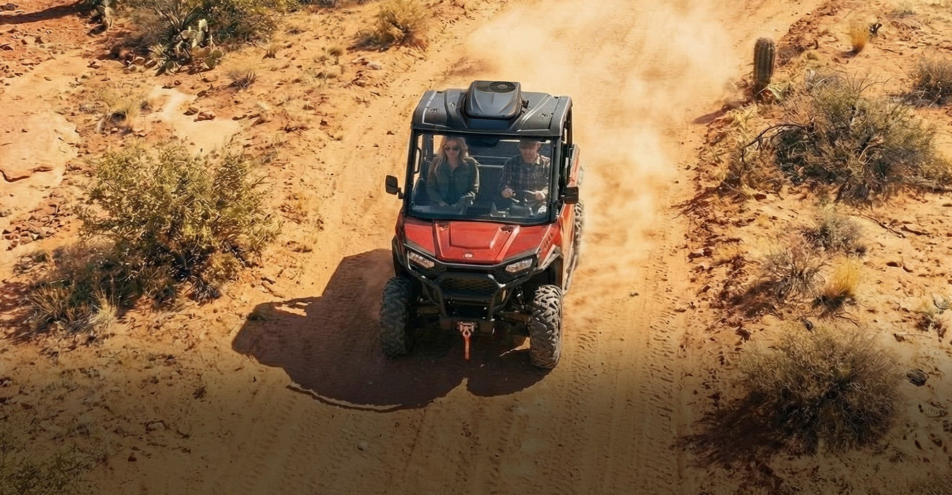 Red off-road UTV driving on a dusty dirt trail through sparse desert brush.