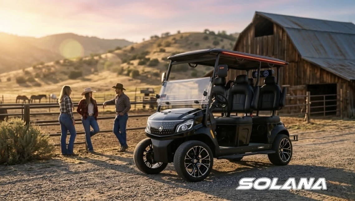 Three people beside a black utility cart near a rustic barn at sunset in a desert landscape