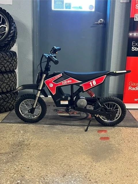 Small black-and-red dirt bike parked indoors beside a gray door and stacked tires.