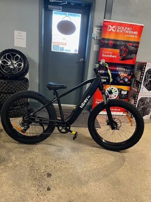 Black fat-tire bicycle displayed indoors beside stacked product boxes and a glass door.
