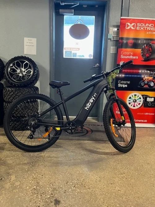 Black NCM electric mountain bike indoors beside stacked wheels and a red display stand