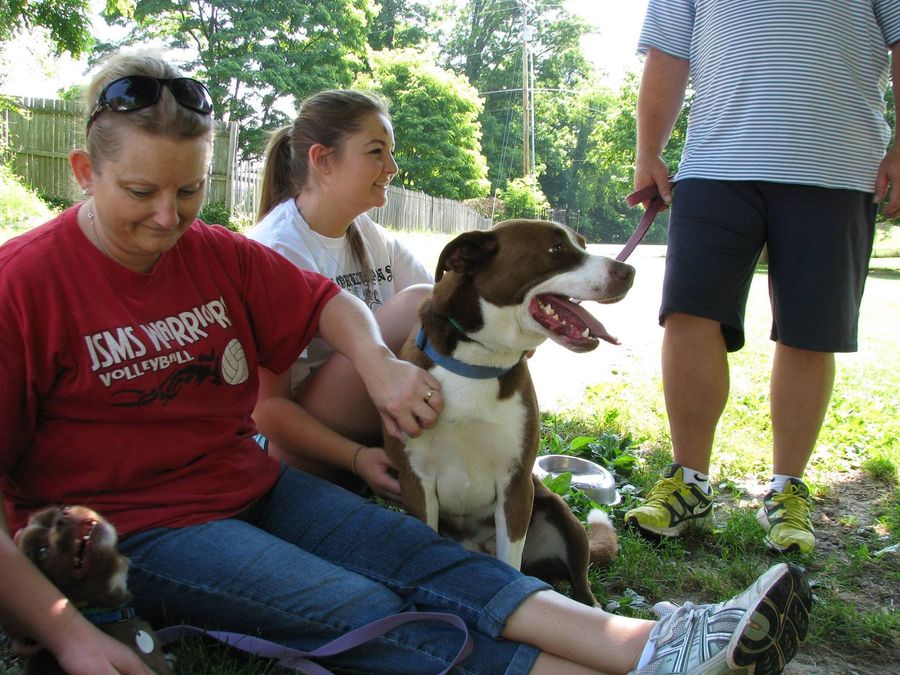 Woman sitting with a dog outdoors while another person stands nearby in a garden.