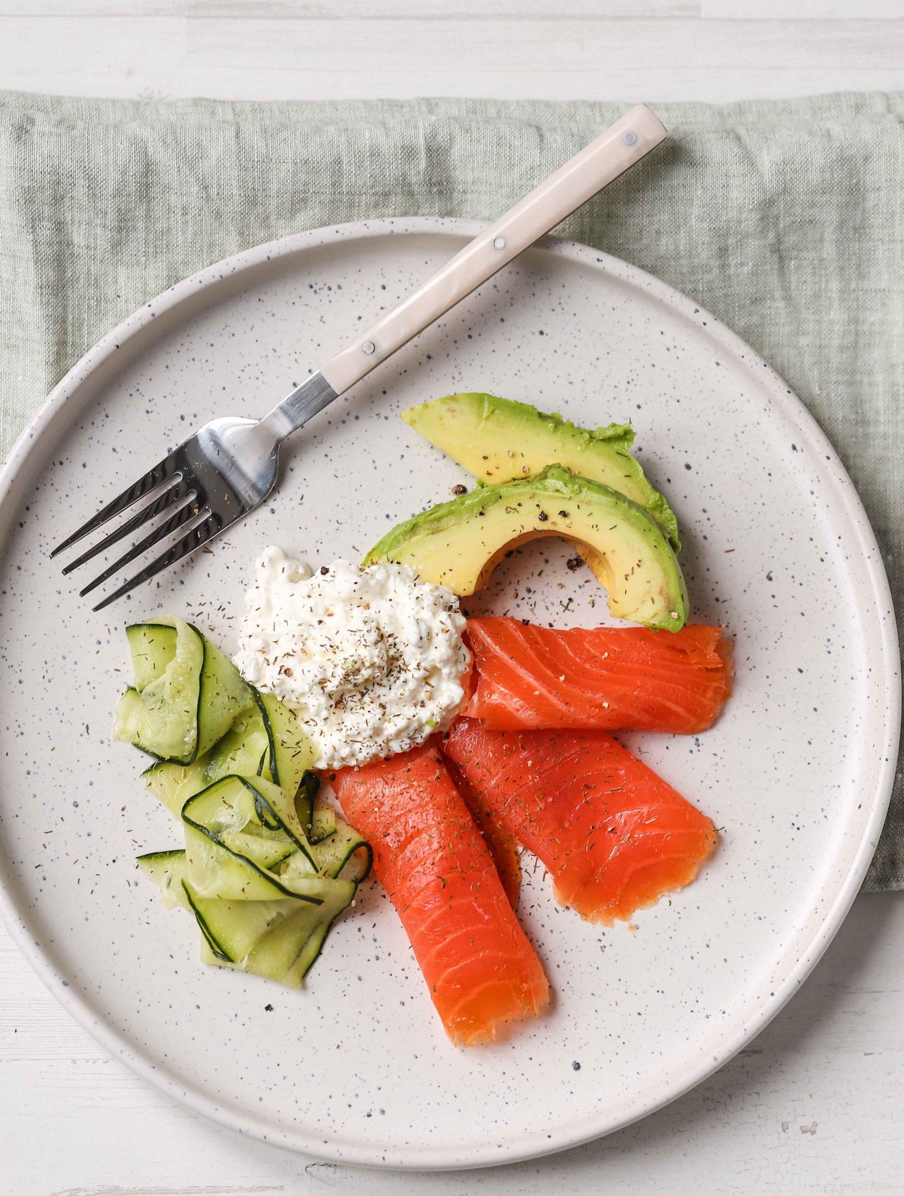 A white plate topped with smoked salmon , avocado , and cucumber with a fork.