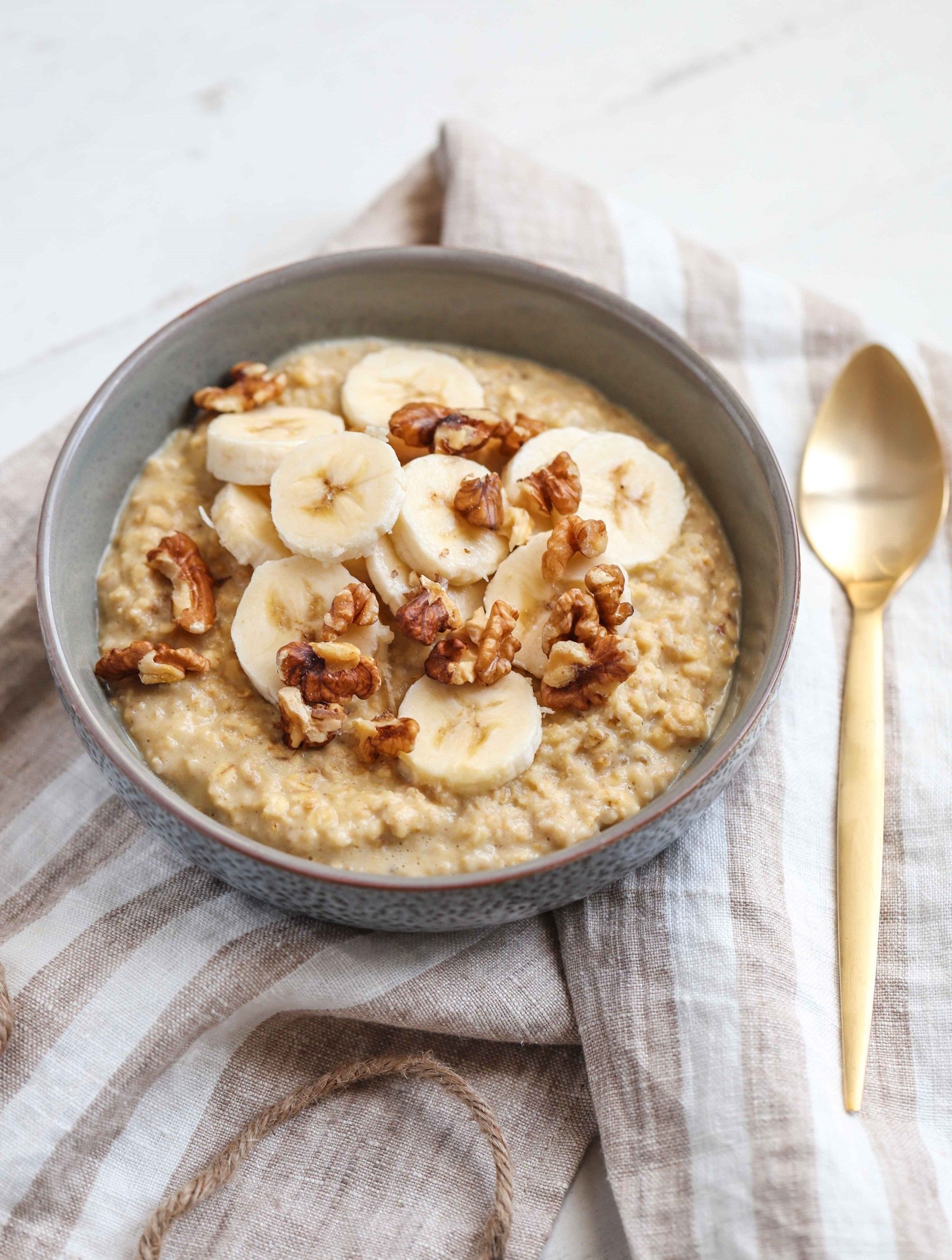 A bowl of oatmeal with bananas and walnuts next to a gold spoon.