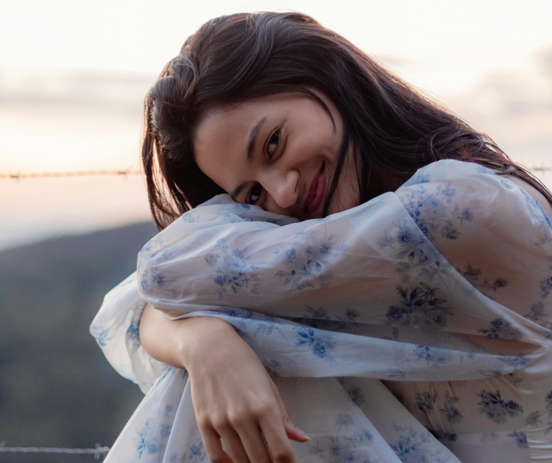 Crouched on the beach, close up shot of a teen in a long sleeve dressed, smiling into the camera.