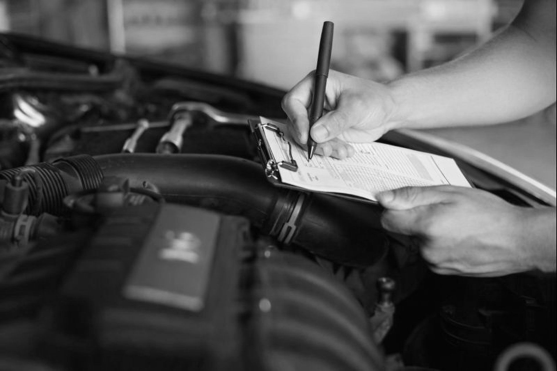 A mechanic writes on a clipboard while inspecting an open car engine in a garage.