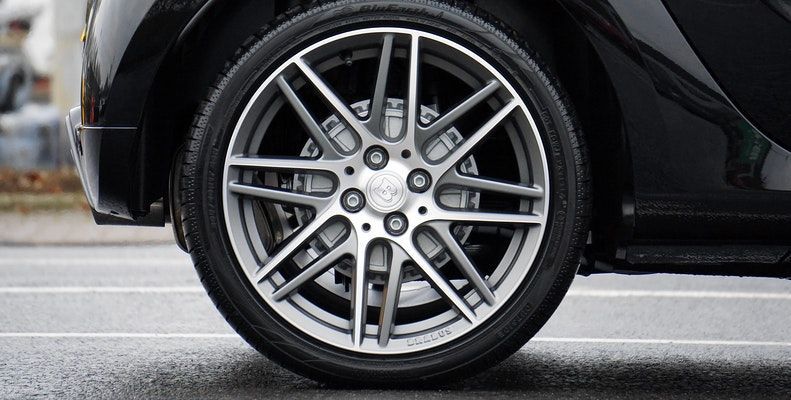 A close-up of a black car's multi-spoke silver alloy wheel on a paved surface.
