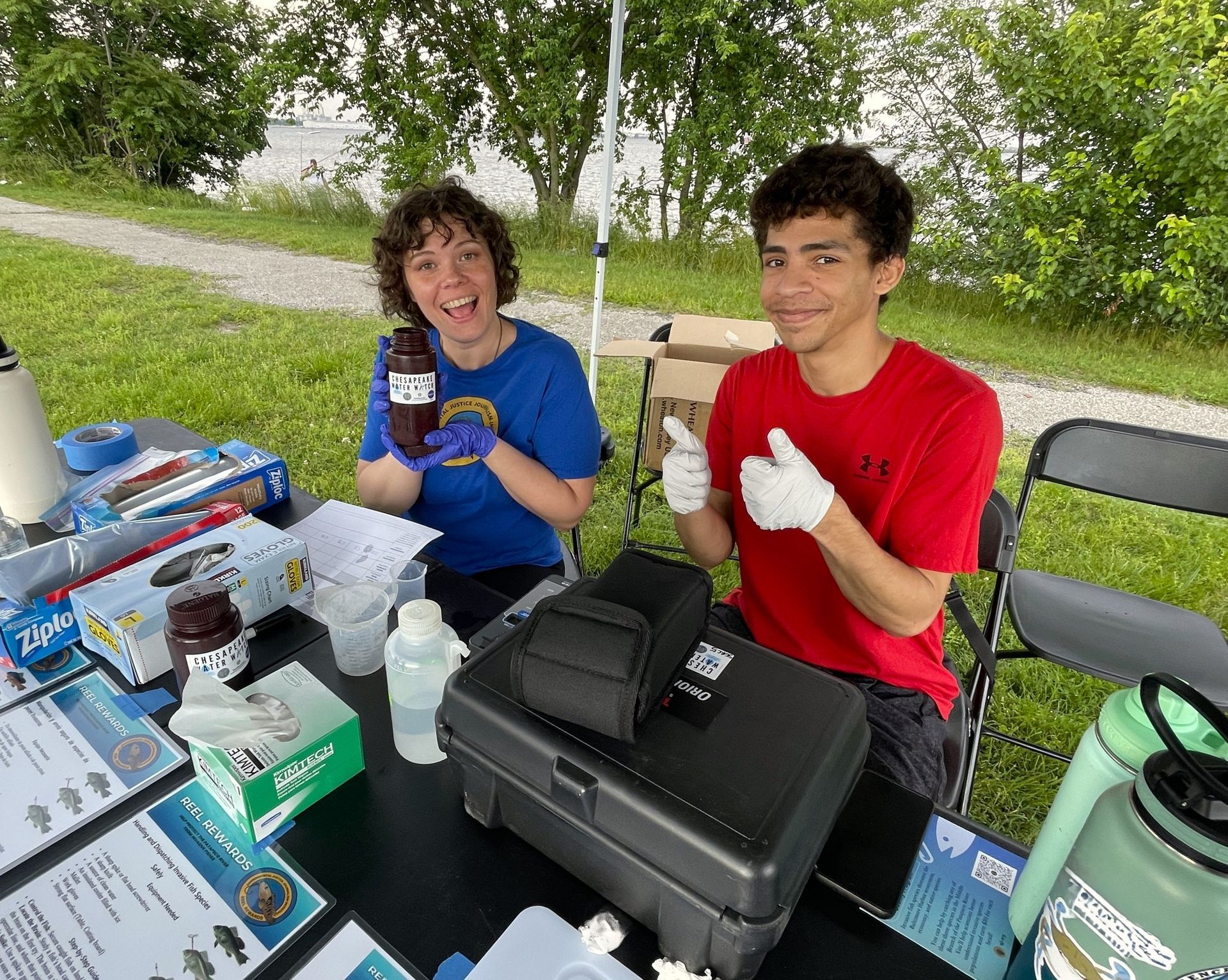 A woman on the left in a blue shirt is holding up a brown bottle that says Chesapeake Water Watch on it. She is smiling. The man to her right is smiling and giving a thumbs up. The background is of plants and the water.