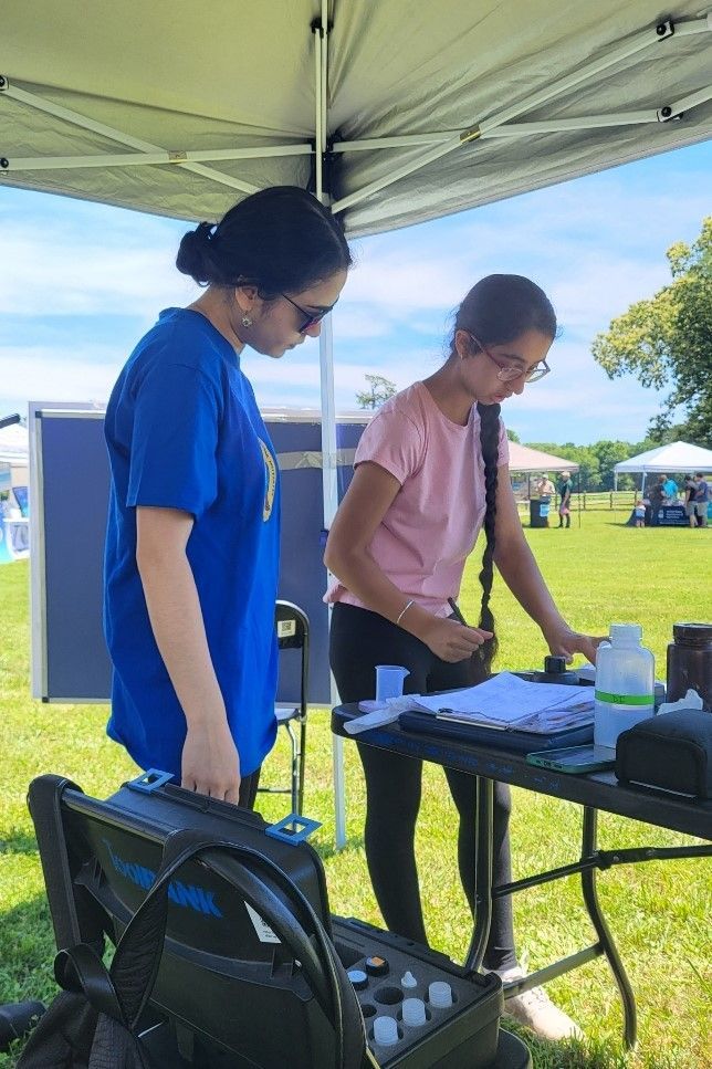 Two women look down at a table with data sheets and water samples. They are standing under a tent in a nature area.