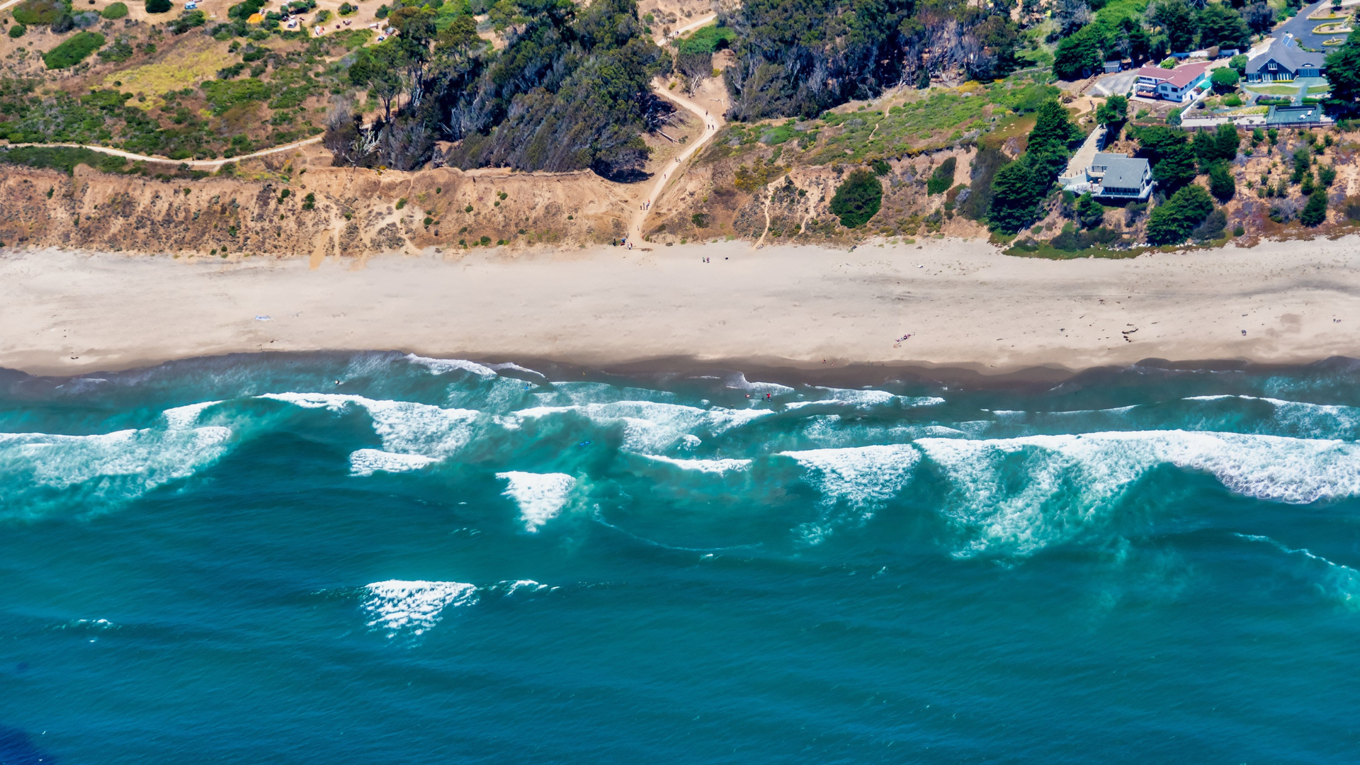 Aerial view of a beach with turquoise waves crashing onto the sandy shore, cliffs, and houses in the background.