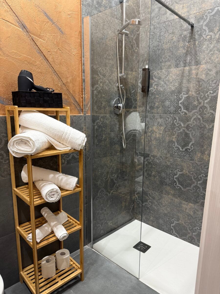 Doccia con porta in vetro, pareti piastrellate, mensola in legno con asciugamani e asciugacapelli.