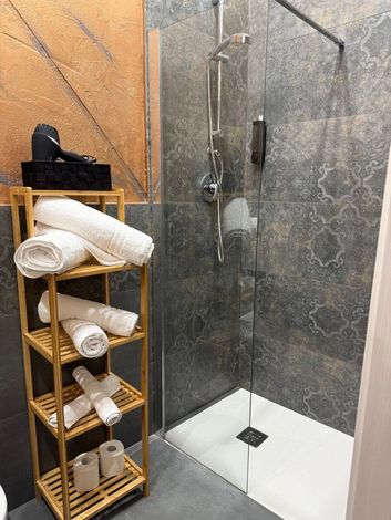 Doccia con porta in vetro, pareti piastrellate, mensola in legno con asciugamani e asciugacapelli.