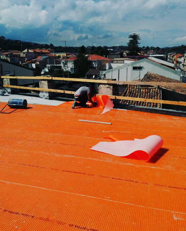 Persona che stende del materiale di copertura arancione su un tetto, con uno sfondo di paesaggio urbano.