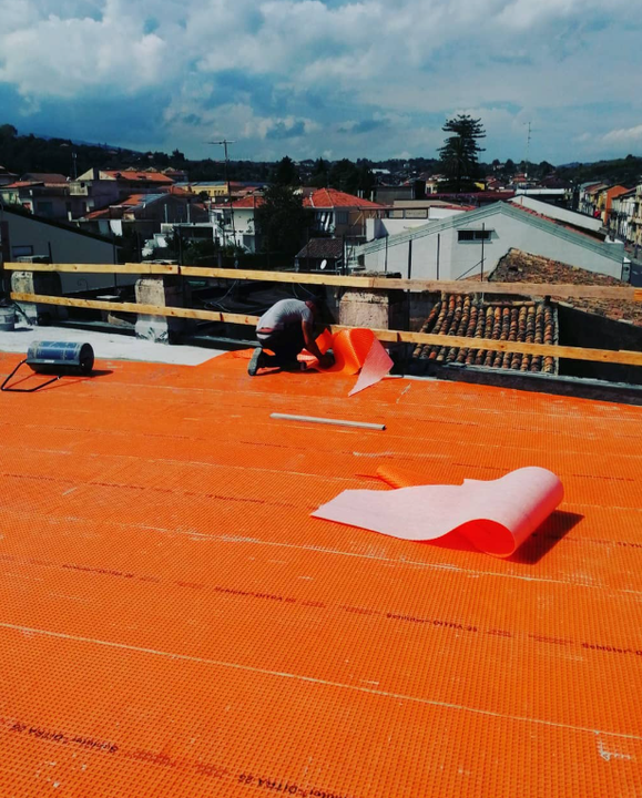 Un operaio edile installa del materiale di copertura arancione su un tetto, con edifici cittadini sullo sfondo.