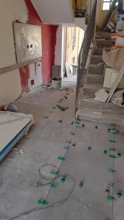 Cantiere interno. Posa delle piastrelle del pavimento, con distanziatori. Sono visibili il muro rosso, le scale e i detriti.
