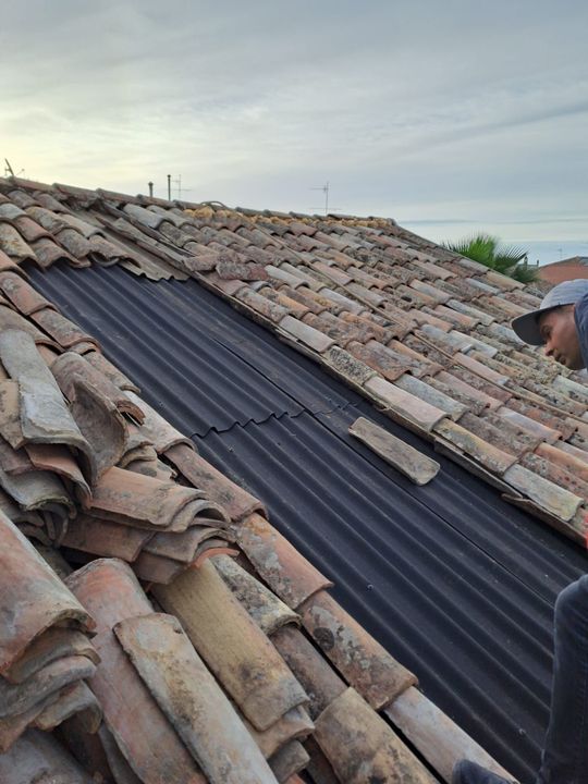 Un operaio sta sostituendo le tegole su un tetto in tegole di argilla, con visibile il sottofondo ondulato nero. Cielo nuvoloso.