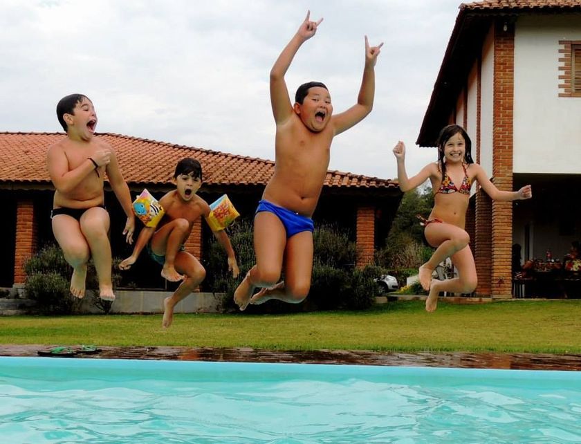 Crianças pulando em uma piscina no quintal, com uma casa e gramado ao fundo.