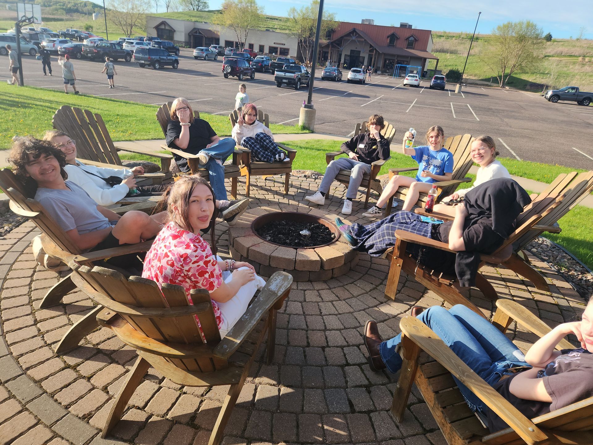 Group of teens around a fire pit, socializing. Sunny day, outdoor setting.