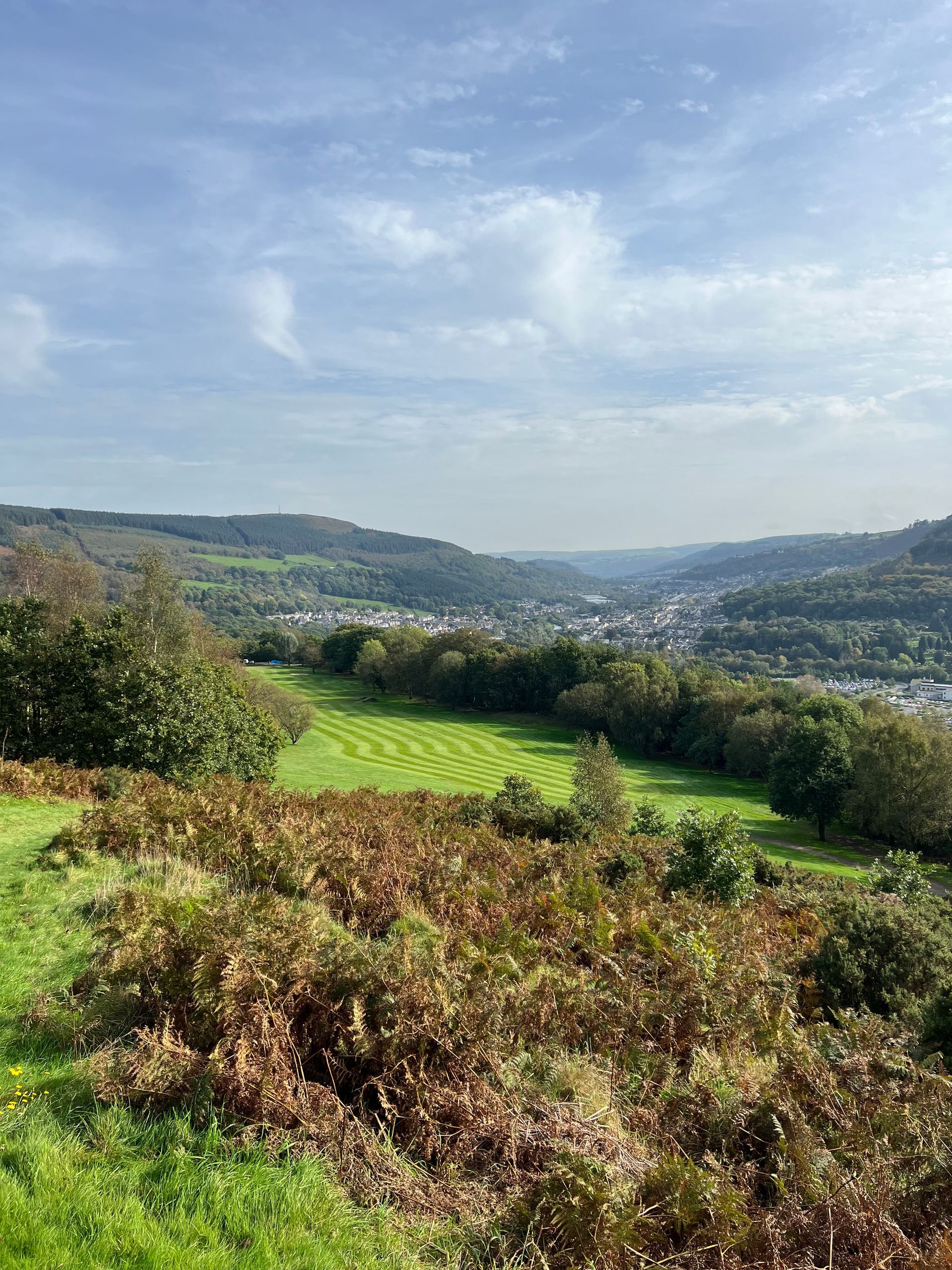 A valley landscape with a golf course in the center, surrounded by hills and a town in the distance under a cloudy sky.