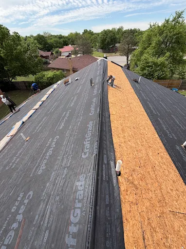 Roof under construction with black underlayment, orange plywood, and workers against a suburban backdrop