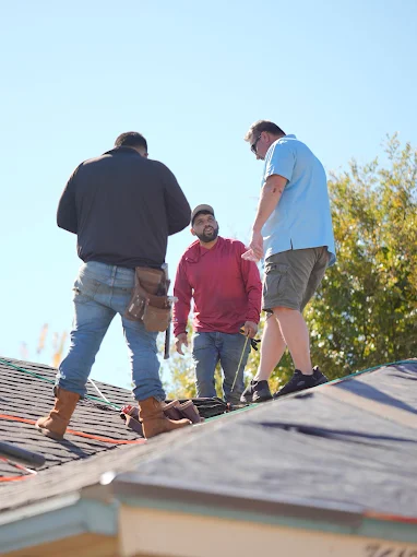 Three workers standing on a roof, inspecting shingles under a clear blue sky.