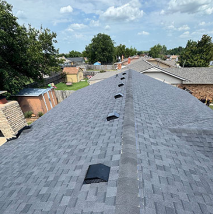 Roofing work on a gray shingle roof, viewed along the ridge with vents and neighboring houses visible.