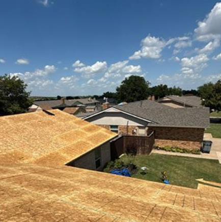 Roofing work on a suburban house under a bright blue sky, with nearby homes and a backyard visible