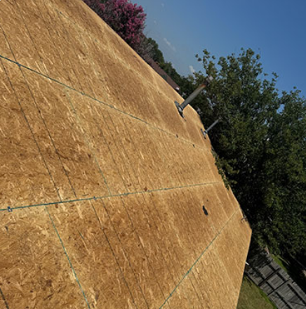 Tilted plywood wall with a small pipe or fixture, outdoors beside trees under a blue sky