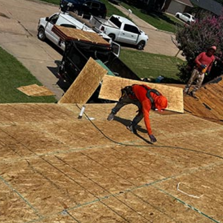 Worker in orange gear nailing plywood roofing on a house under construction, with trucks nearby.