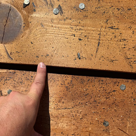 Finger pointing at a gap between two weathered wooden boards with nails and saw marks.