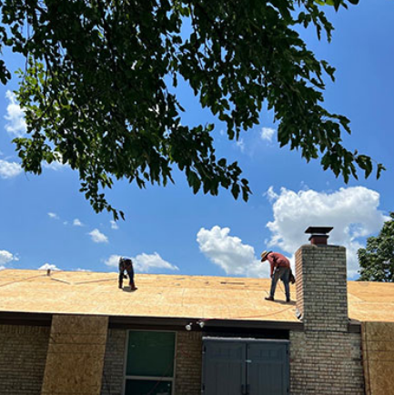 Two workers on a house roof under a blue sky, installing shingles beside a brick chimney.