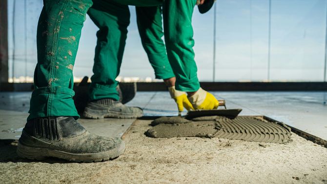 Worker in green pants and yellow gloves smoothing wet concrete with a trowel indoors.
