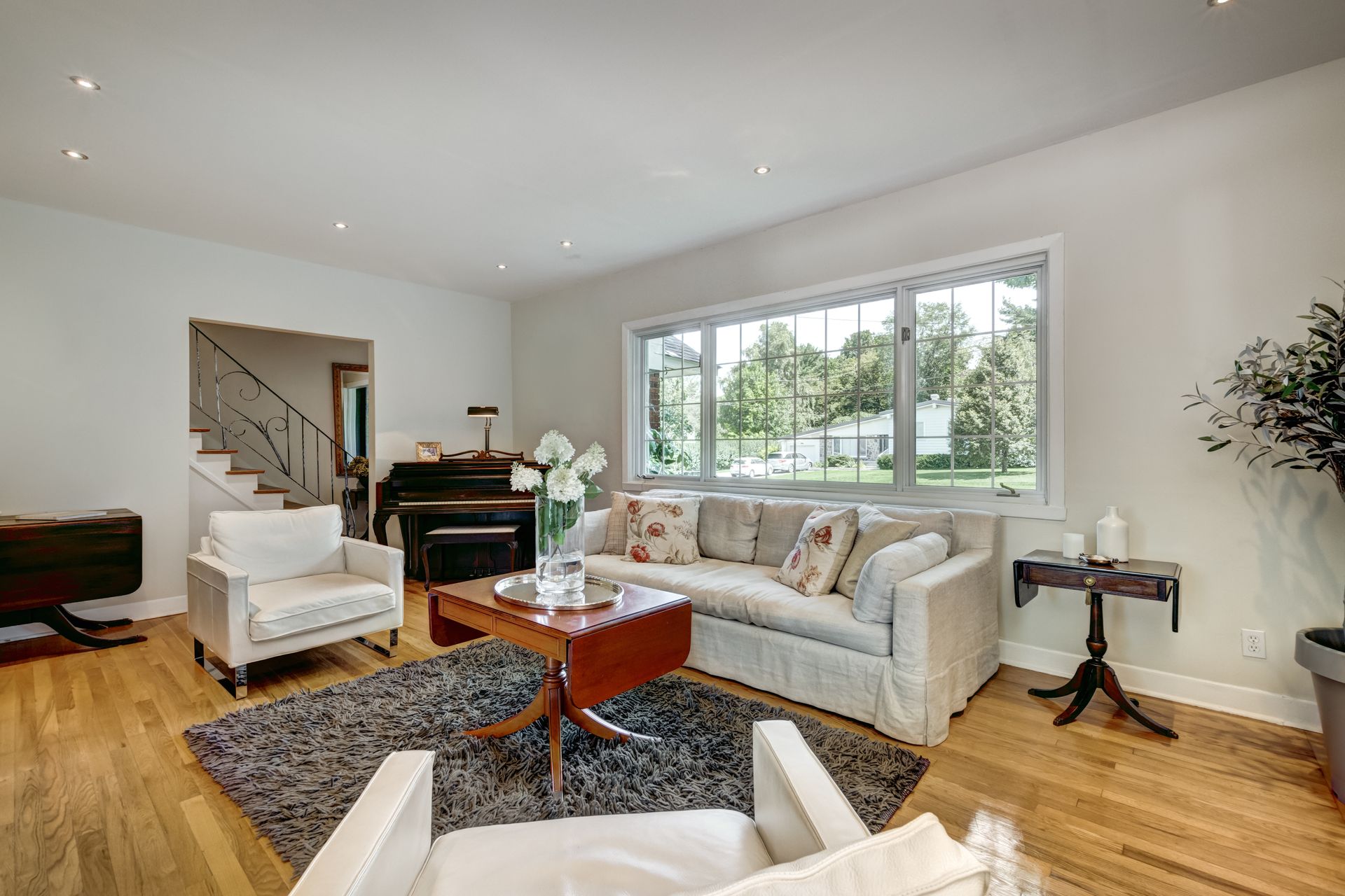 Bright living room with white sofa, armchairs, piano, patterned rug, hardwood floors, and large window views