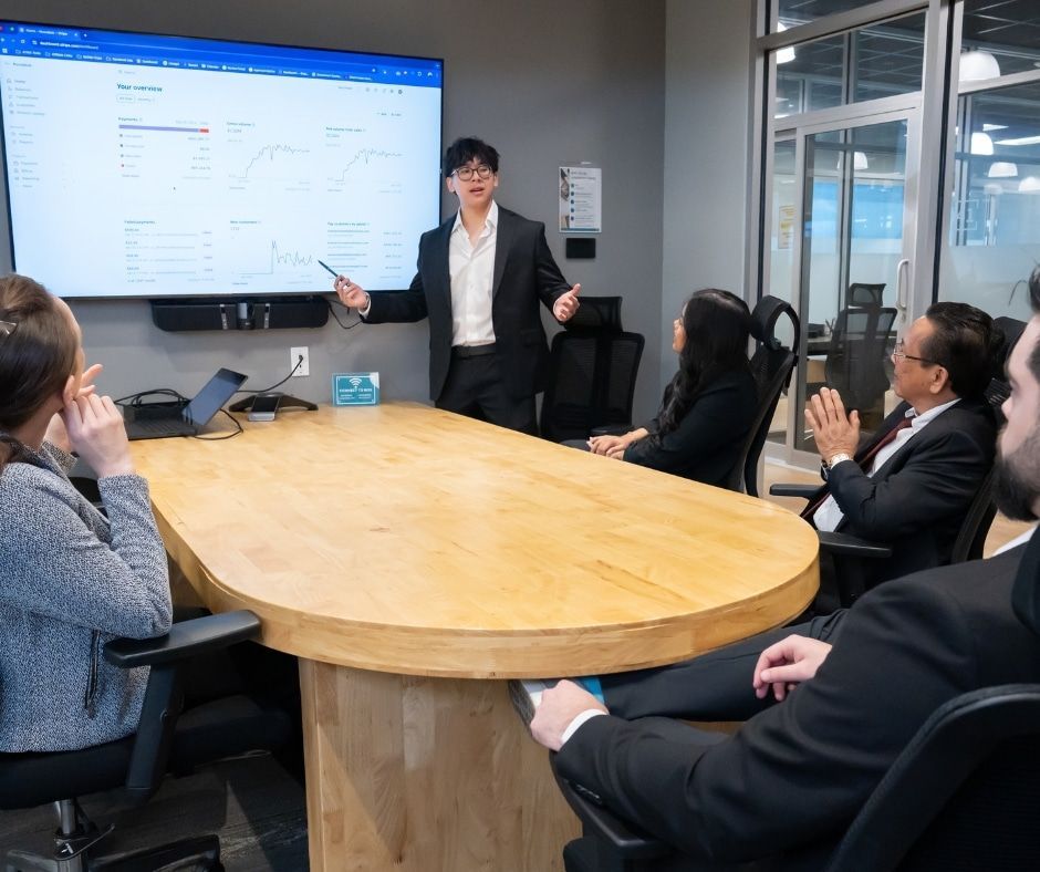 Business meeting in a conference room, with a presenter speaking beside a large screen and attendees seated around a table.
