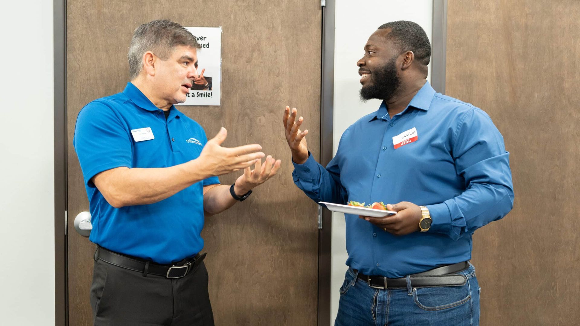 Two coworkers in blue shirts talk in a hallway, one holding a clipboard and the other gesturing.