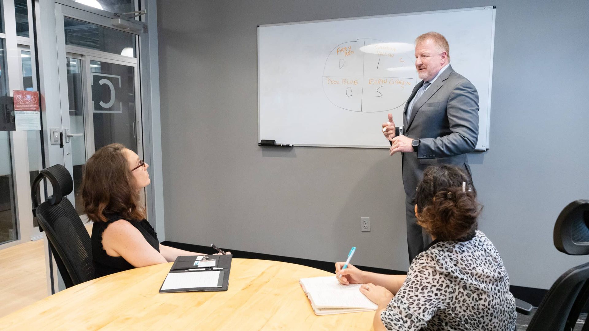 Business meeting in a conference room, with a man presenting at a whiteboard to two seated attendees.