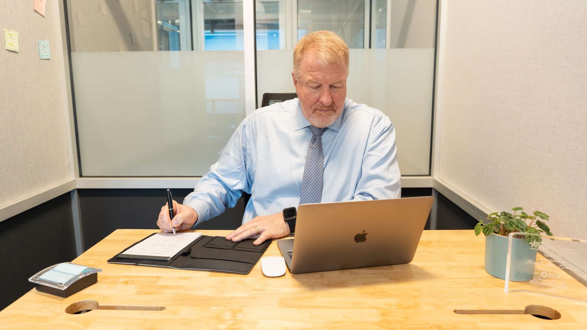 Person working at a desk with a laptop and papers in a small office, writing by hand