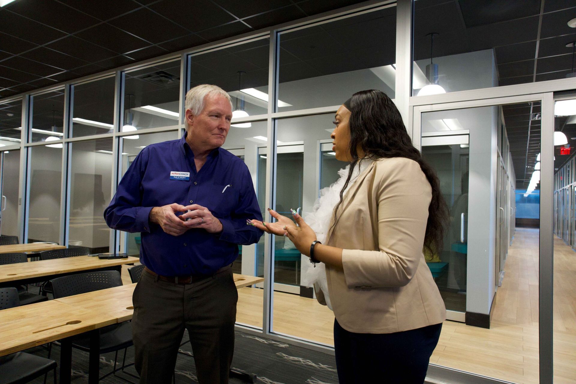 Two people talking in a glass-walled office, one in a blue shirt and one in a beige blazer.